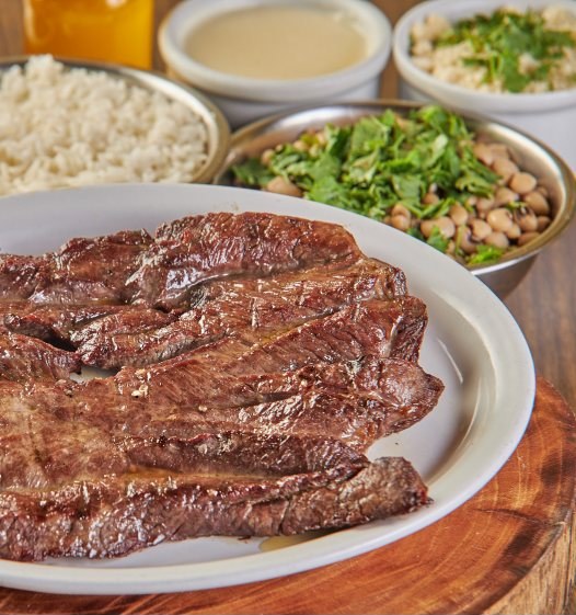 Carne de sol fatiada num prato branco, com arroz, feijão verde, macaxeira e nata.